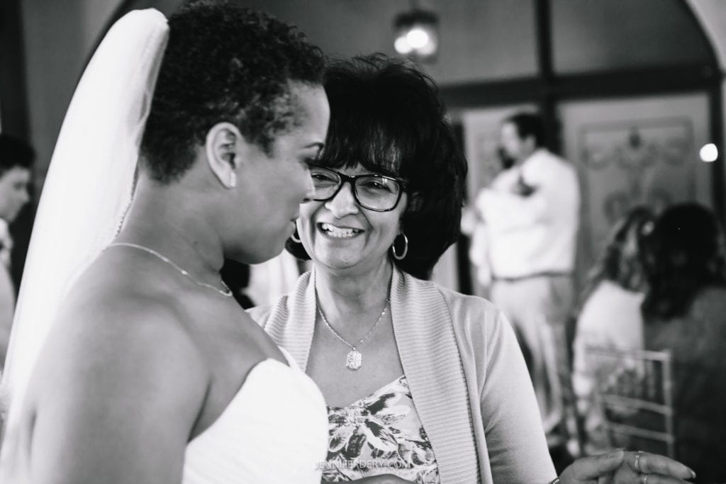 A bride in a strapless wedding dress and veil talks with an her mother wearing glasses and a light sweater. They are smiling and appear to be sharing a special moment at the Balboa Park wedding reception, with other guests visible in the background.