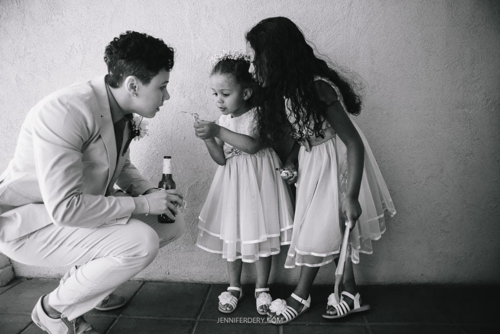 A person in a light-colored suit leans down to interact with two children in dresses. One child is holding a bubble wand and seems focused, while the other child looks on, holding a small toy. The setting appears to be indoors against a plain wall, reminiscent of preparations for a Balboa Park wedding.