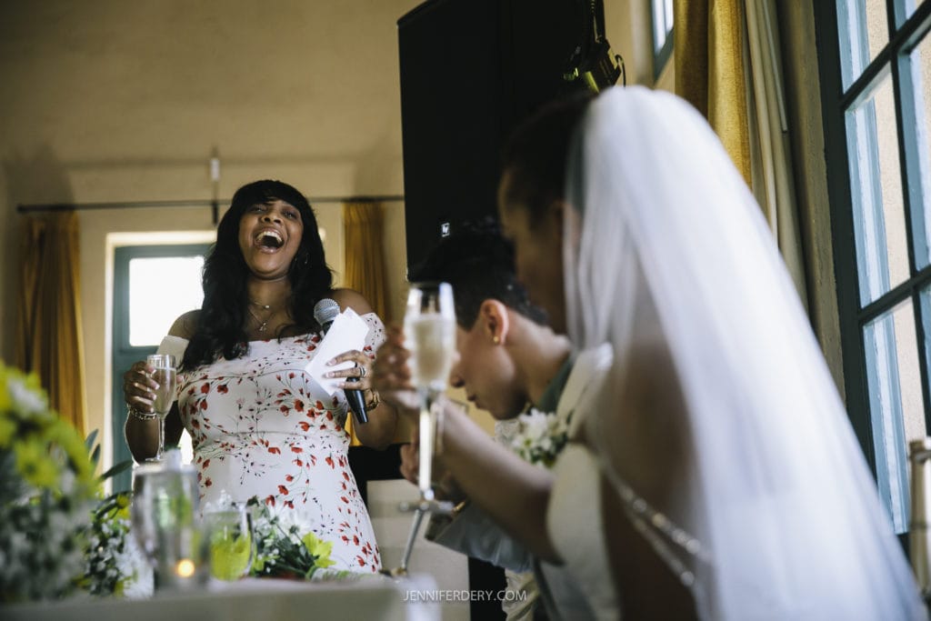 At a Balboa Park wedding, a woman in a floral dress is giving a toast with a microphone and laughing. In the foreground, the bride in her white gown and veil reacts joyfully with a drink. Another seated guest is partially visible between them, adding to the festive atmosphere of the celebration.