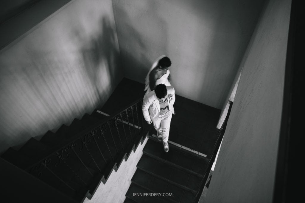 A black and white photo capturing a couple walking up a staircase at their Balboa Park wedding. The elegant railing and contrast between light and shadow create a dramatic atmosphere. The man is slightly ahead, and both are dressed formally. Photo credit: jenniferdery.com.
