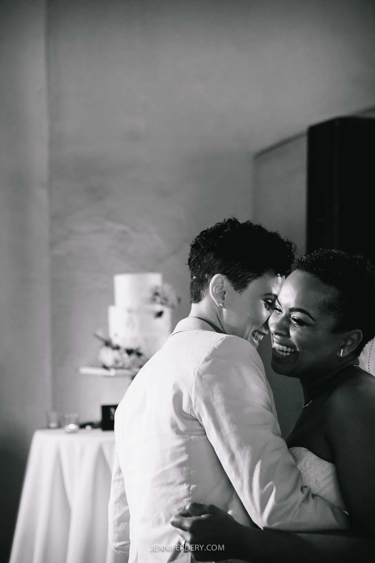 A black and white photo of two people embracing and smiling, at a Balboa Park wedding reception. One is wearing a light-colored suit and the other a strapless dress. They are in front of a table with a tiered wedding cake on it, slightly out of focus in the background.