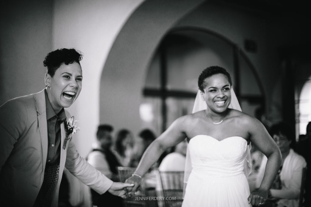 A black and white photo of two people holding hands and smiling joyfully at their Balboa Park wedding. The person on the left is dressed in a suit, and the person on the right is wearing a wedding dress and veil. The background shows an archway with guests seated.