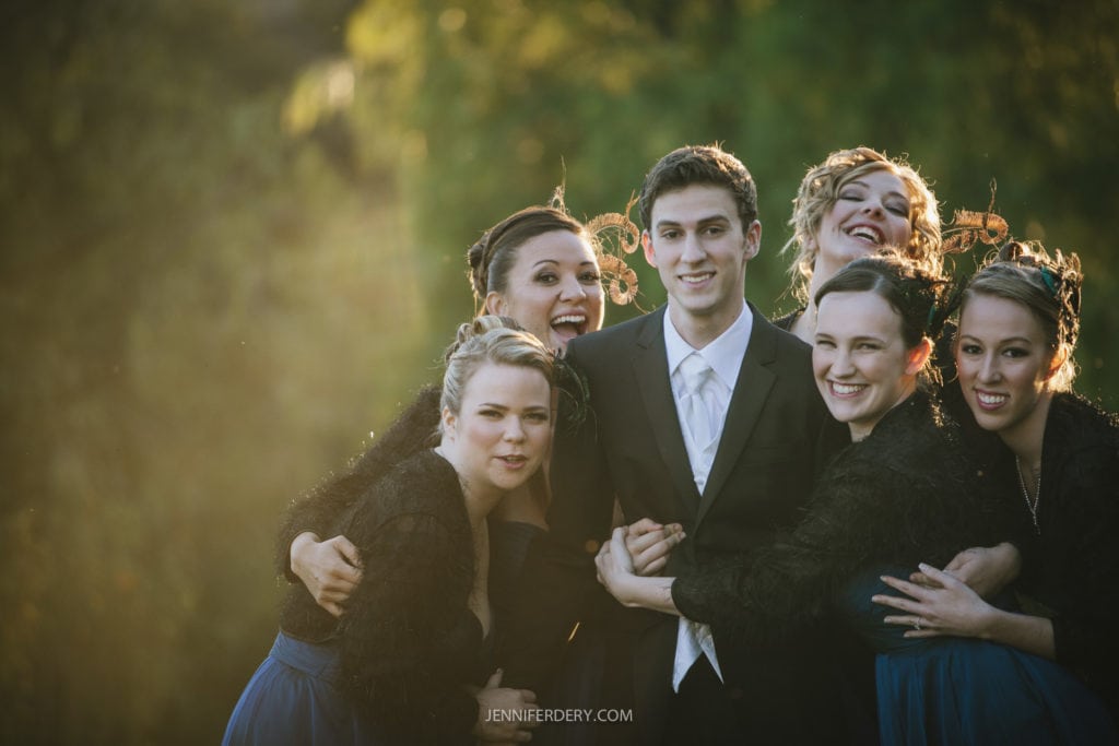 A groom in a suit stands outdoors surrounded by five smiling women in matching dark dresses with fur accents. The group is huddled closely together against a backdrop of lush greenery. Warm sunlight filters through the trees, creating a soft, golden glow.