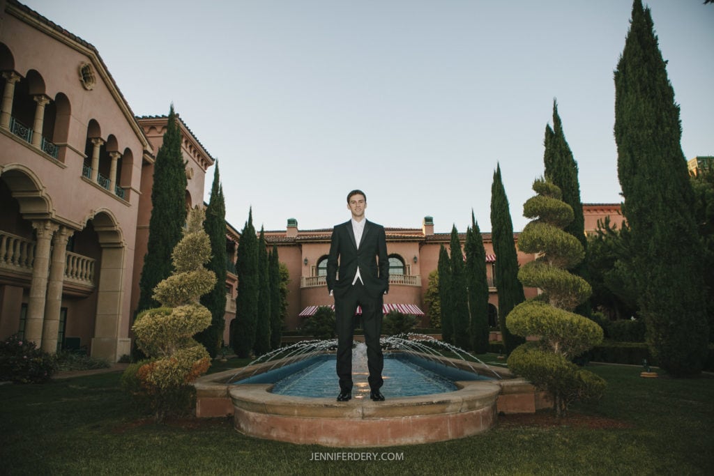 A man in a black suit stands confidently on a circular stone platform in front of a long, narrow fountain. He is positioned between two tall, ornate buildings with neatly trimmed trees and hedges lining the courtyard. The sky is clear and blue.