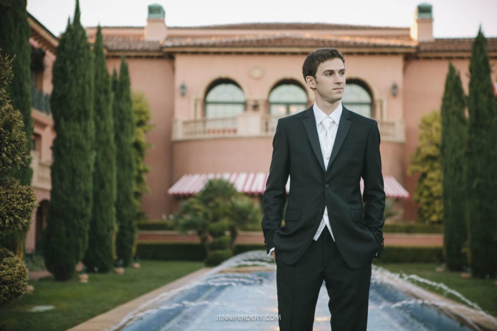 A young man in a black suit and white shirt stands in front of a grand building with tall, narrow trees on either side and a long, narrow fountain in front of him. The building has large arched windows and a tiled roof. The sky above is clear.