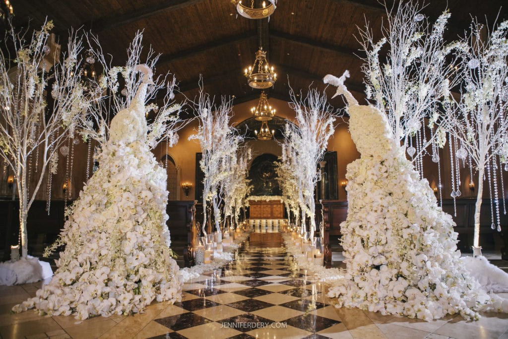 the wedding chapel at the grand del mar with a polished checkered floor is lavishly decorated for a wedding, featuring tall floral arrangements of white peacocks made of flowers and branches. Golden chandeliers hang from the vaulted wooden ceiling, illuminating the elegant and opulent setting.
