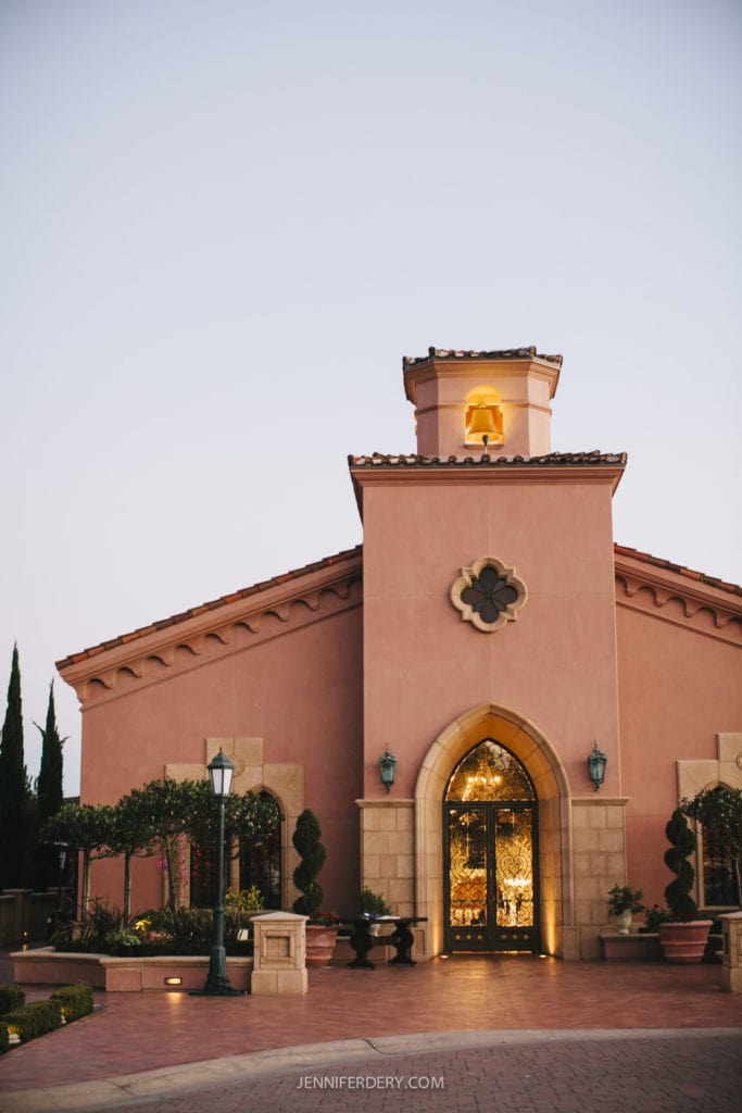 the charming Mediterranean-style chapel building a the grand del mar at dusk, featuring warm terracotta walls, a central arched entrance with glass doors, and a small tower with an illuminated window. Neatly trimmed topiaries and classical street lamps adorn the entrance.