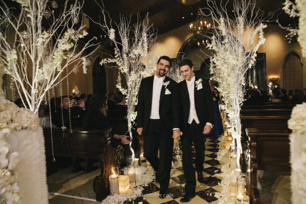 Two grooms in tuxedos walk hand-in-hand down an elegant, candle-lit aisle adorned with white flowers and decorative branches. They are smiling, surrounded by seated guests inside a dimly-lit, ornate venue, celebrating their wedding ceremony.