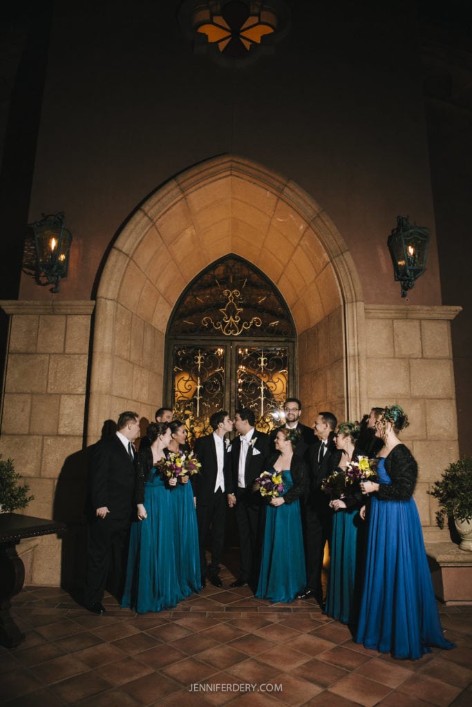 A wedding party at the grand del mar stands outside an elegant, arched doorway at night. The bride and groom share a kiss in the center while their bridesmaids in teal dresses and groomsmen in black suits surround them, smiling and looking on. The scene is warmly lit.