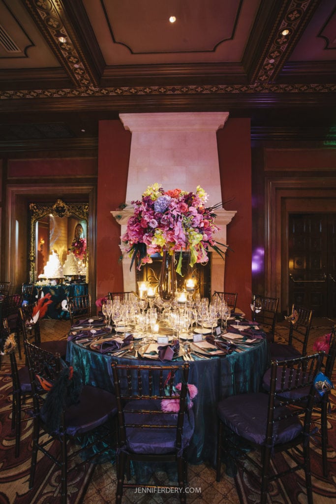 A wedding decorated round table set for formal dining, featuring elaborate floral centerpieces with vibrant pink, purple, and blue flowers. The table is elegantly laid out with glassware and cutlery, and is situated in front of a grand fireplace in an ornately decorated room.