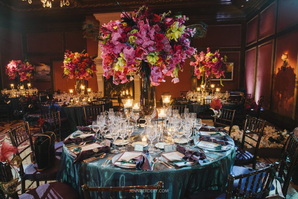 An elegant banquet hall featuring round tables set with luxurious tableware, glasses, and ornate floral centerpieces with pink and yellow flowers. The dimly lit room has a warm ambiance, with candles adding a soft glow to the sophisticated setting.