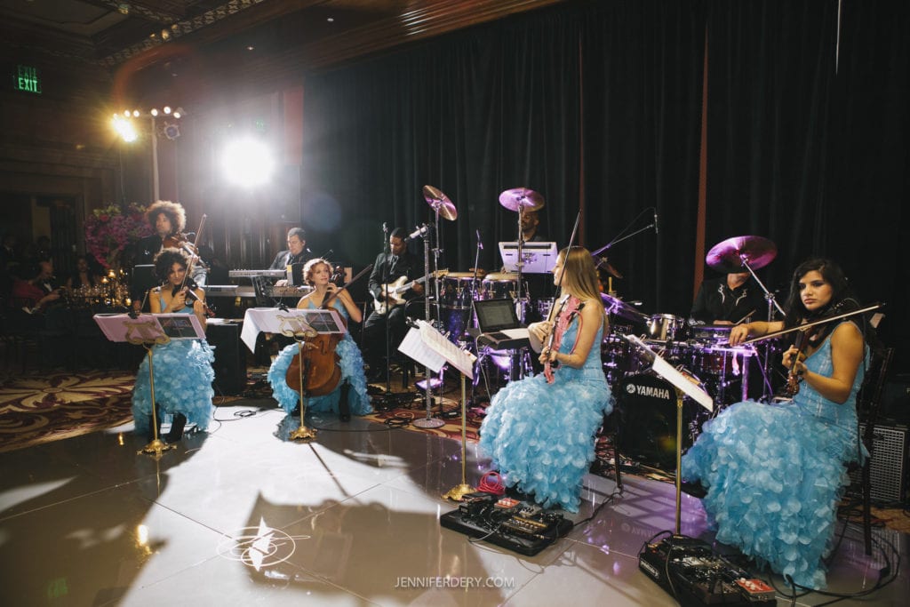 A musical ensemble in blue dresses performs on stage with various instruments including violins, a cello, clarinets, and percussion. The background features dark curtains and stage lights. The performers are focused and the atmosphere is elegant.