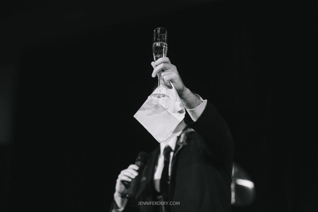 A person in a suit and tie holds up a glass award and an envelope while standing on a stage. The person's face is obscured by the raised objects. They appear to be holding a microphone in their other hand.