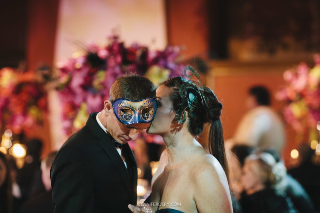 A couple dressed elegantly in formal attire shares an intimate moment at a masquerade ball. Both are wearing decorative masks, with the man in a suit and the woman in a strapless dress. The background features vibrant, colorful floral arrangements and dim lighting.