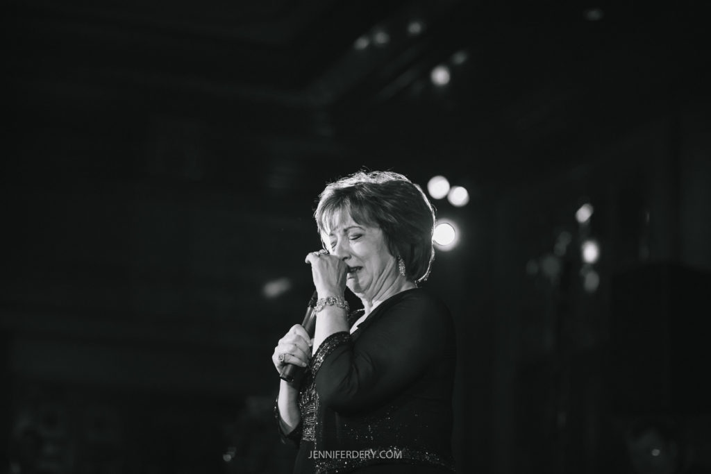 A black and white photograph of a person holding a microphone with their right hand and smiling slightly. They appear to be in the middle of a wedding toast on stage, illuminated by stage lights. The background is dark and out of focus.