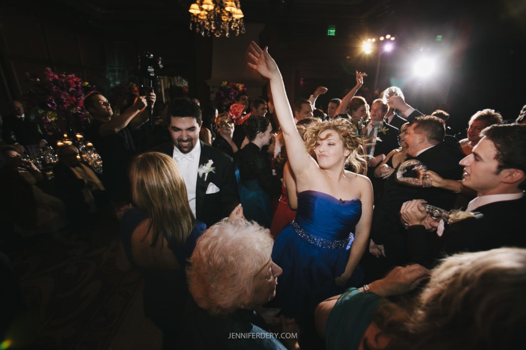 A lively wedding reception with guests dancing energetically. One woman in a blue dress is in the center with her arm raised. Others are smiling, clapping, and moving to the music. The room is decorated with flowers, and lights create a festive atmosphere.