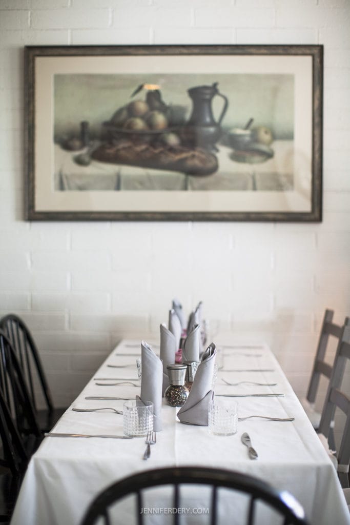 A long dining table set for a meal at Little Lion Cafe with white tablecloth, grey napkins, silverware, and glasses. Black chairs surround the table. A framed still life painting of bread, fruits, and a jug hangs on a white brick wall above the table.