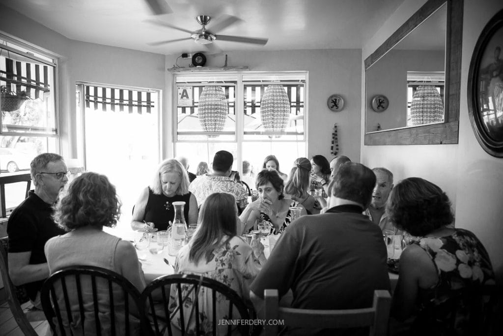 A group of people sit around a large table at Little Lion Cafe, engaging in conversation. The black and white photo captures the intimate atmosphere, with light filtering through the windows and ceiling fans overhead. A large mirror hangs on the wall.