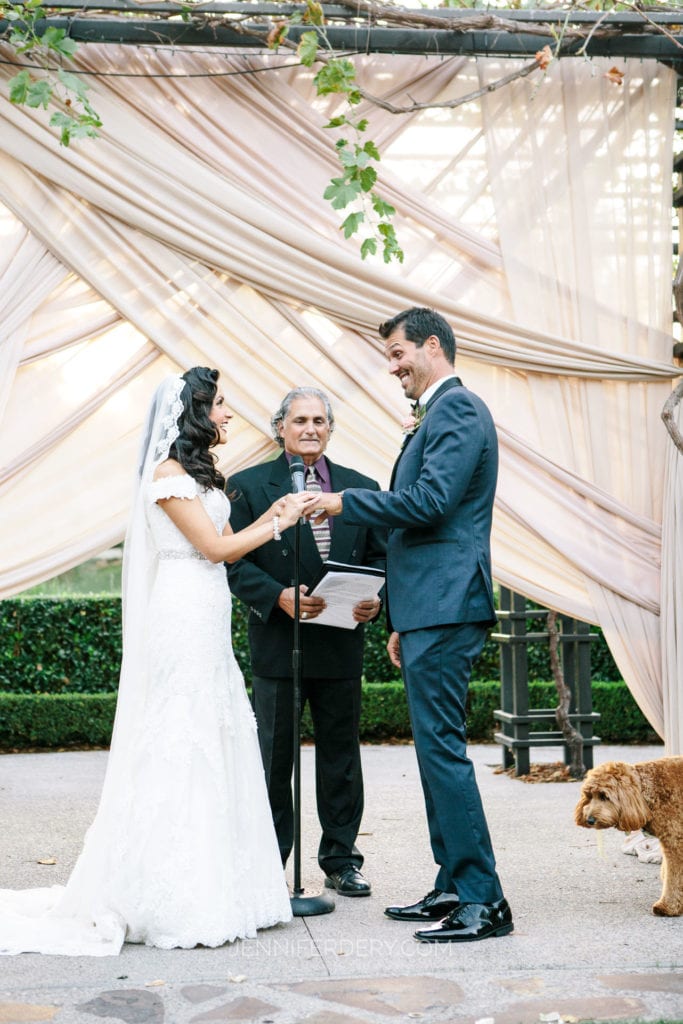 A bride and groom stand facing each other, holding hands, in front of an officiant during an outdoor wedding ceremony at Rancho Bernardo Inn. The bride wears a white dress and veil, while the groom is in a dark suit. A small dog is seen near the groom's feet. The background features draped fabric and greenery.