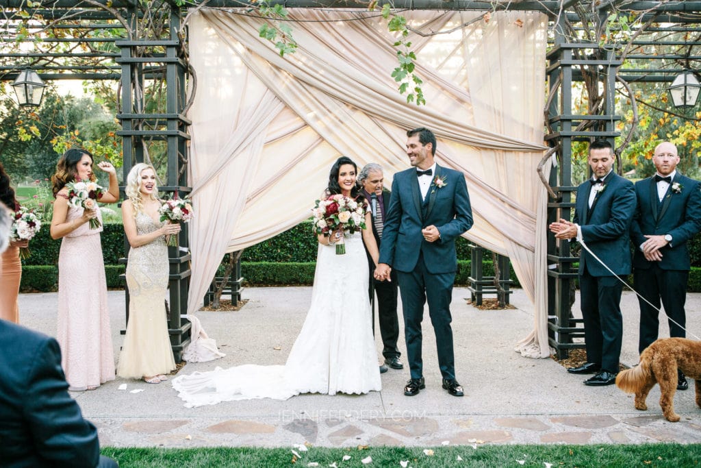 A newlywed couple stands hand in hand under a pergola adorned with fabric and vines at Rancho Bernardo Inn. Guests, including the wedding party, surround them. A flower girl plays with a small dog on the right. The bride holds a bouquet and wears a white dress; the groom is in a dark suit.