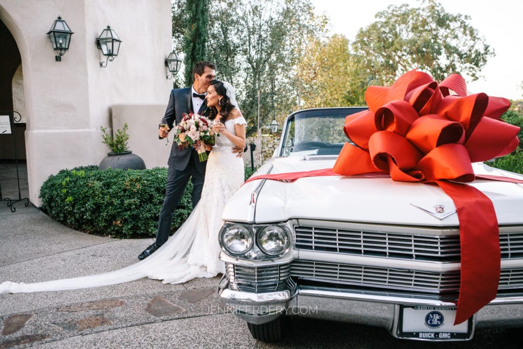 A bride and groom stand close together, smiling, as they admire a bouquet of flowers. The scene evokes a touching Mother's Day portrait, with a vintage white car adorned with a large red bow nearby. The setting appears elegant, framed by a white building and lush greenery in the background.