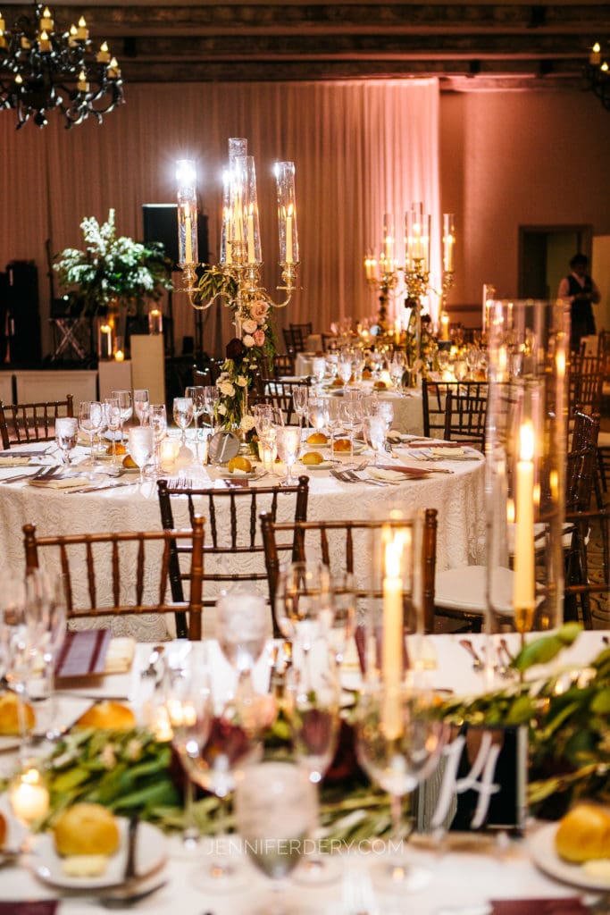 An elegant, dimly lit wedding reception at Rancho Bernardo Inn features round tables adorned with white tablecloths, tall floral centerpieces, and glowing candles. Elegant place settings include wine glasses, plates, and bread rolls. The ambiance is warm and inviting.