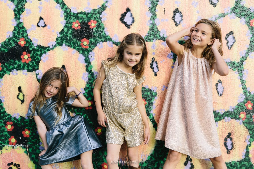Three young girls stand in front of a colorful, floral-patterned wall. The girl on the left wears a metallic blue dress and poses with her hands on her head. The middle girl is in a sparkling gold outfit, and the girl on the right wears a shiny, peach-colored dress. All are smiling.