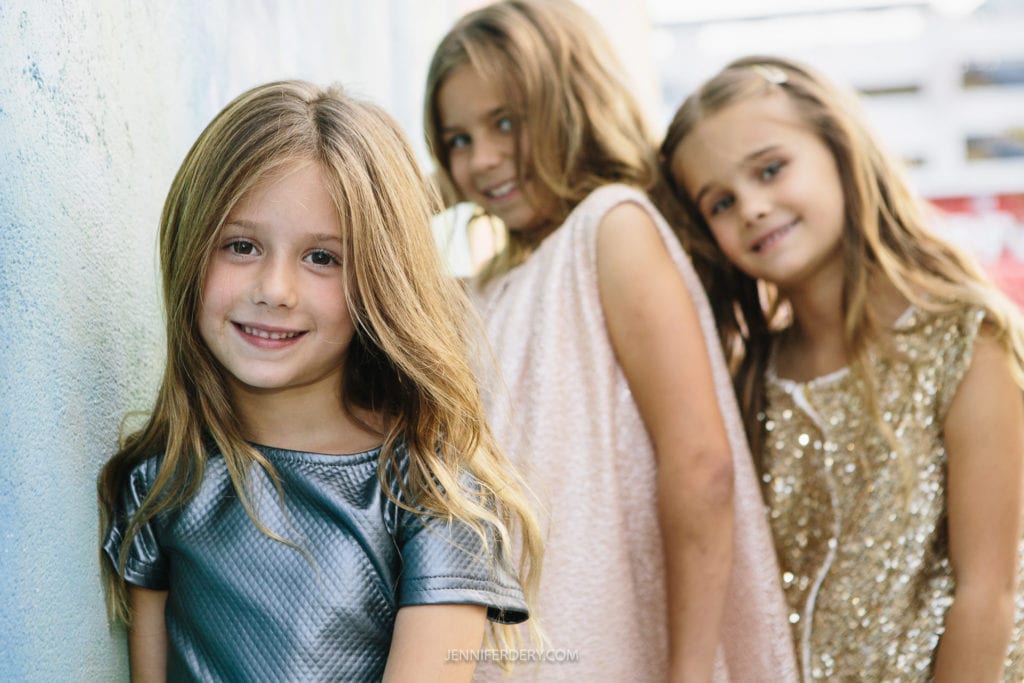 Three young girls with long hair stand closely together, smiling at the camera. The girl in the foreground wears a silver dress, while the two girls behind her wear a light pink dress and a gold dress. They stand against a wall, creating a cheerful and friendly scene.