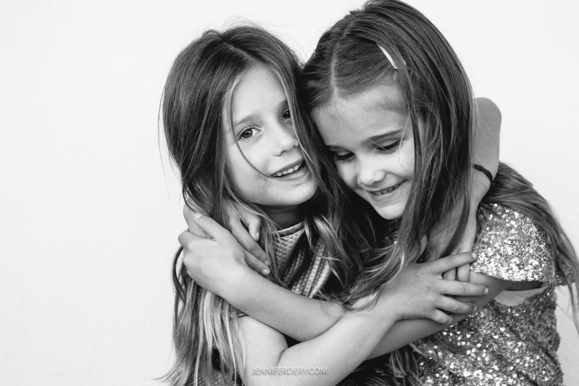Two young girls embracing and smiling at each other in a black and white photo. They have long hair and are showing signs of joy and friendship.