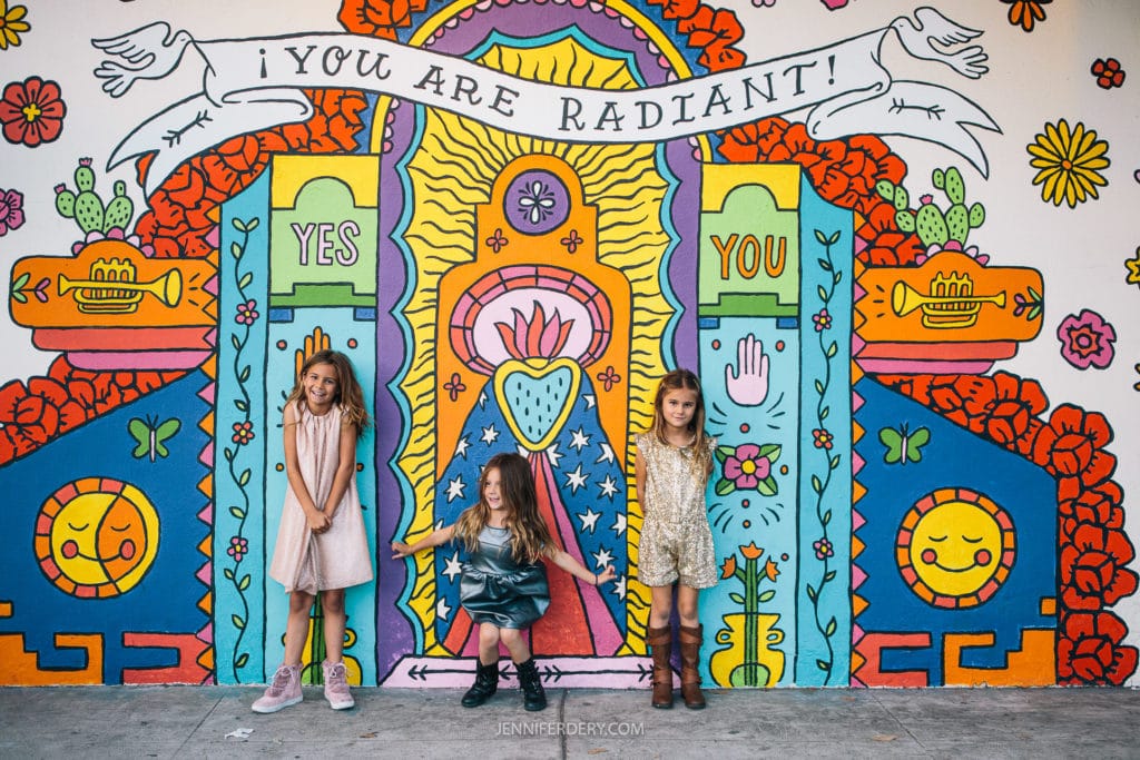 Three children stand in front of a colorful mural with vibrant designs and the words "YOU ARE RADIANT" above a central heart motif. The mural features flowers, stars, and the words "YES" and "YOU" in bold letters.