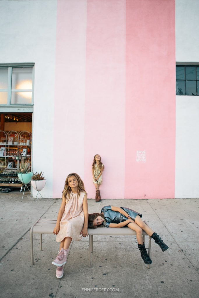 Three young girls pose in front of a building with a large pink stripe. One girl stands in the foreground leaning against a bench, another lies on the bench, and the third stands in the background. The scene captures a playful and stylish moment.