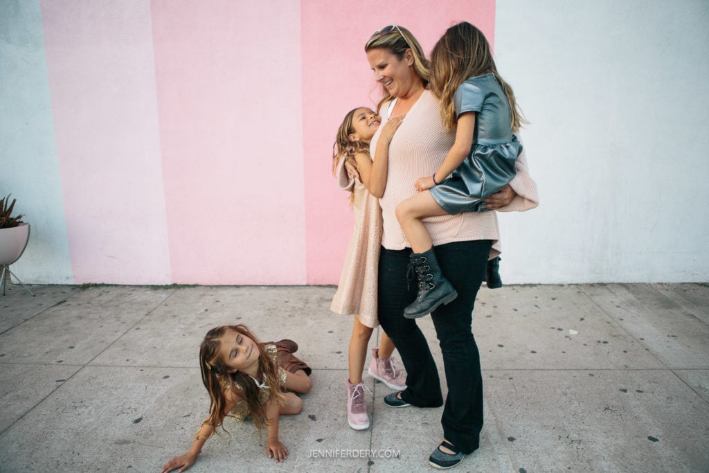 A woman smiles while carrying a young girl in a blue dress on her left arm. Another girl in a pink dress hugs her from the front. A third girl in a beige dress sits on the ground, looking up. They are in front of a pastel pink and blue wall.