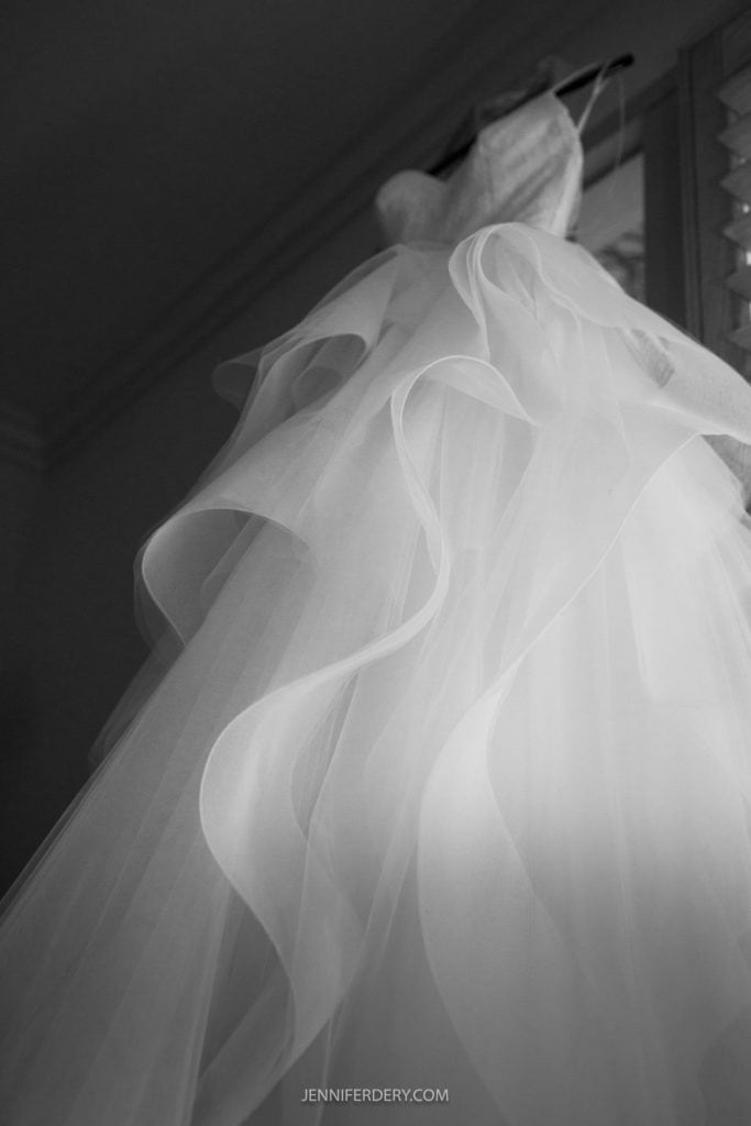A black and white photo of a flowing, layered bridal gown From White Floral hanging on a hanger. The intricate design features multiple ruffled layers of tulle, creating a cascading effect. Captured atRancho Bernardo Inn Photos, the low angle highlights the delicate texture and soft details of the dress.