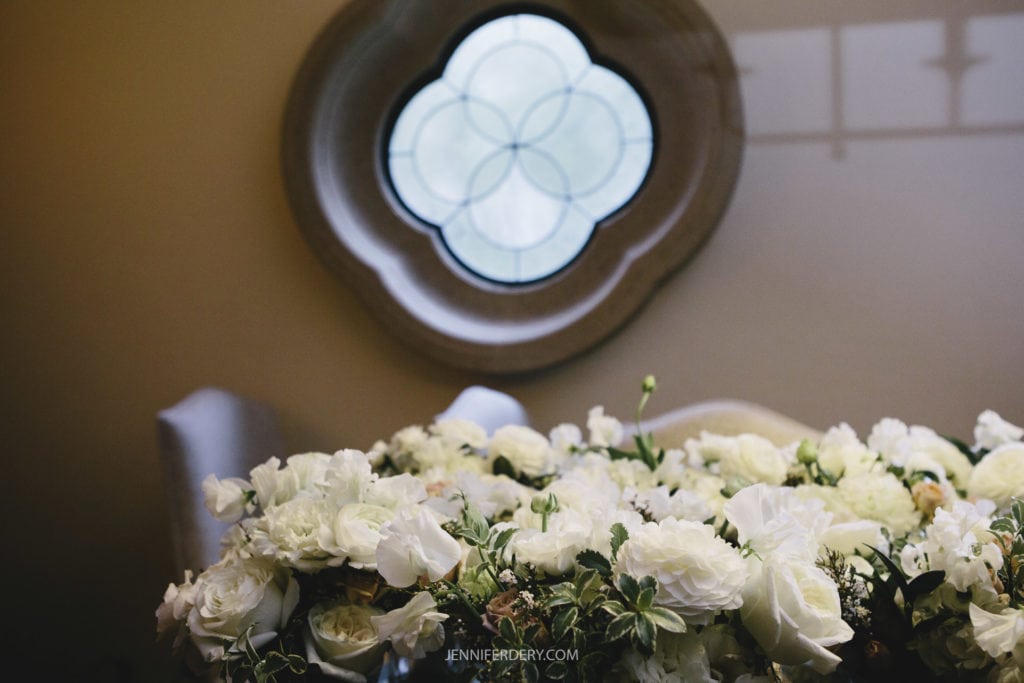 A room with a round decorative window features a large bouquet of white roses and peonies in the foreground. The window, reminiscent of Rancho Bernardo Inn wedding Photos, has a flower-like pattern of overlapping circles, and the walls are softly lit, creating a peaceful ambiance.