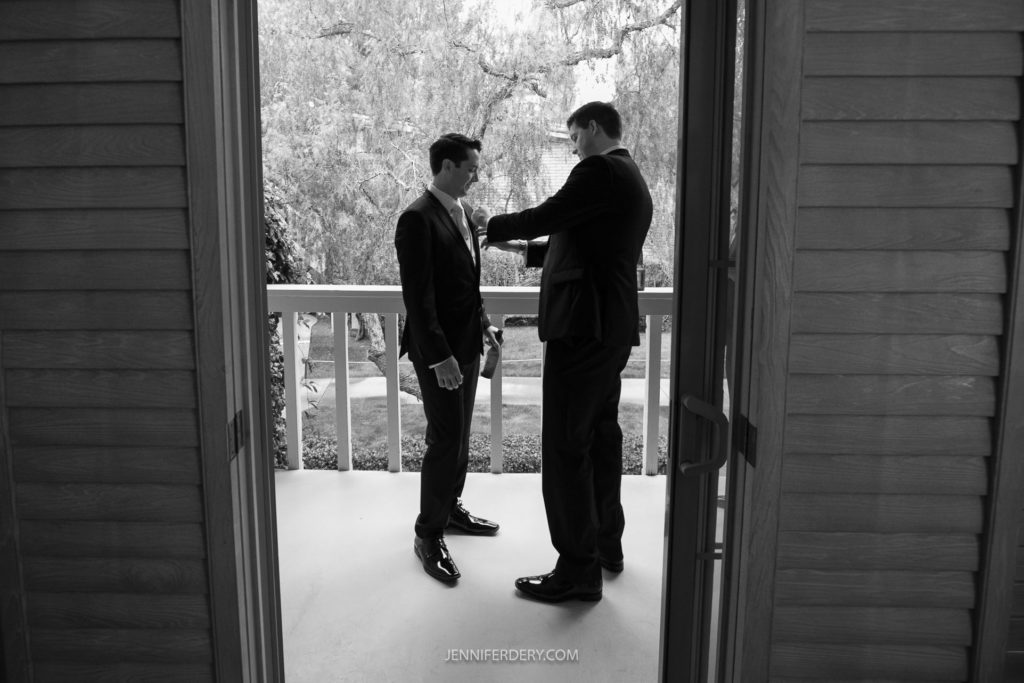 Black and white photo of two men on a balcony, one helping the other adjust his tie. Both are dressed in suits and standing between an open pair of doors with foliage and trees visible behind them. The watermark "JENNIFERDERY.COM" subtly marked. Reminiscent of timeless Rancho Bernardo Inn