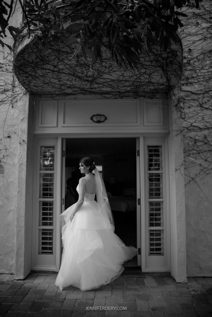 Black and white photo of a bride standing in a doorway under a canopy of vines, looking back over her shoulder and smiling. She is wearing a flowing wedding dress with a long veil. The doorway opens into a room, its ground paved with bricks—an exquisite moment captured at Rancho Bernardo Inn