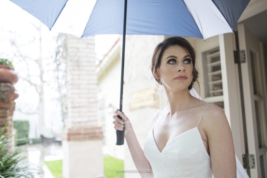 A woman in a white sleeveless wedding dress holds a blue and white umbrella. She looks to her left with a thoughtful expression, reminiscent of Rancho Bernardo Inn wedding photos. In the background, there is a blurred outdoor scene with greenery and a building.