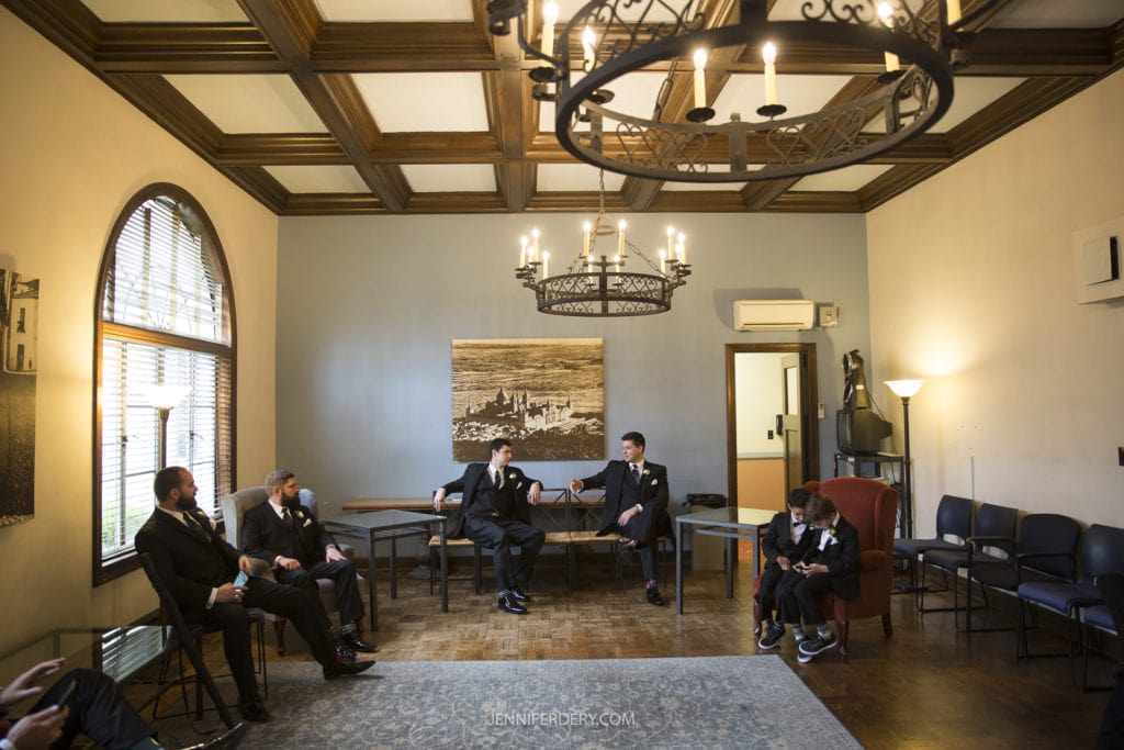 A group of men in suits sit in a formal, elegantly decorated room with wooden ceiling beams and chandelier lighting, reminiscent of Founders Chapel Wedding Photos. Some are engaged in conversation, while others sit or stand quietly. A large window allows natural light in, highlighting the classic ambiance.