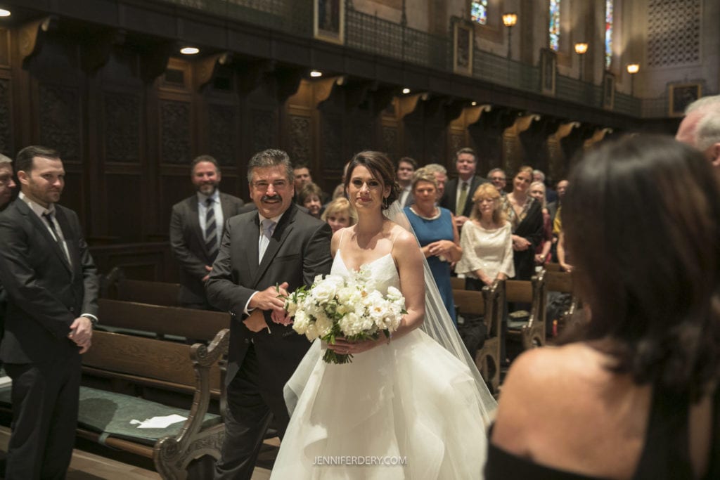 A bride in a white gown holding a bouquet of white flowers walks down the aisle escorted by a man in a tuxedo. Guests stand and smile in the background of the beautifully decorated Founders Chapel, with stained glass windows capturing the moment for unforgettable wedding photos.