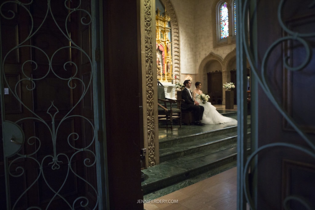 A bride and groom are seated inside the Founders Chapel on a green marble platform, near an altar decorated with golden elements. The scene is viewed through open wooden doors with ornate ironwork, capturing the essence of serene Founders Chapel wedding photos in the softly lit interior.