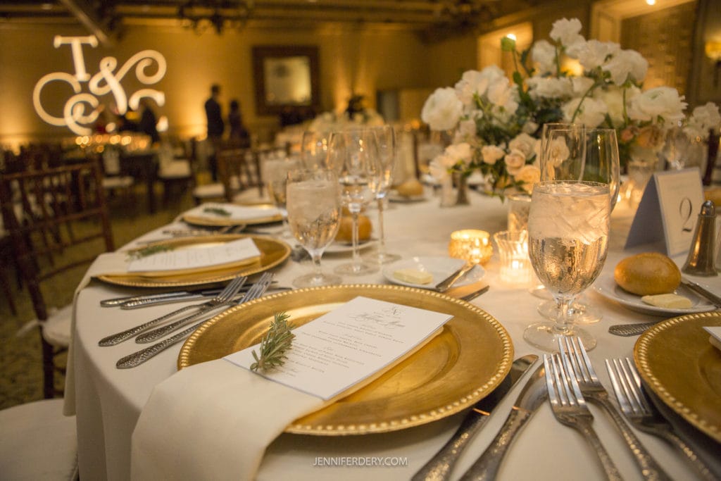 A beautifully set table at a wedding reception, featuring gold-rimmed plates with menus, elegant glassware, and silver cutlery. The centerpiece includes white flowers and candles, with a backdrop of a monogrammed "T & C" illuminated on a distant wall, perfect for Rancho Bernardo Inn wedding photos.