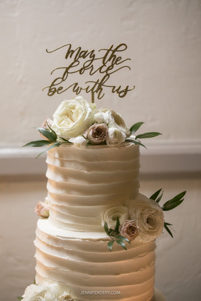 A two-tiered white cake decorated with white and blush roses, greenery, and a gold cake topper that reads, "May the Force be with us." The cake is beautifully showcased against a neutral background, perfect for capturing in your Founders Chapel Wedding Photos.