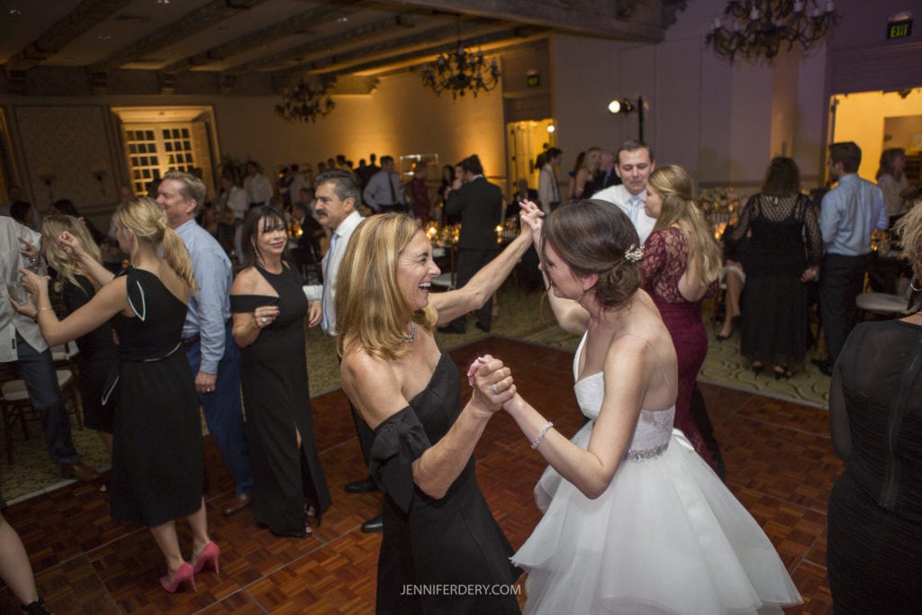 A bride in a white gown joyfully dances with a woman in a black off-the-shoulder dress at a lively indoor wedding reception. Other guests, dressed in formal attire, fill the background, enjoying the celebration. The atmosphere is festive and warm—perfect for capturing unforgettable Founders Chapel Wedding Photos.