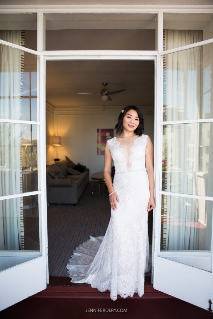 A bride stands in an open doorway at the Japanese Friendship Garden Wedding, smiling and wearing a lacy, floor-length white wedding dress. The room behind her is softly lit and furnished with a couch, lamp, and framed artwork on the wall. The door frame and light drapes add an elegant touch.