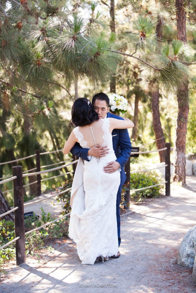 A bride and groom embrace lovingly under tall, lush trees in an outdoor setting at the Japanese Friendship Garden Wedding. The bride is wearing a white lace wedding dress, and the groom is in a dark suit. The surrounding area is scenic with sunlight filtering through the foliage.