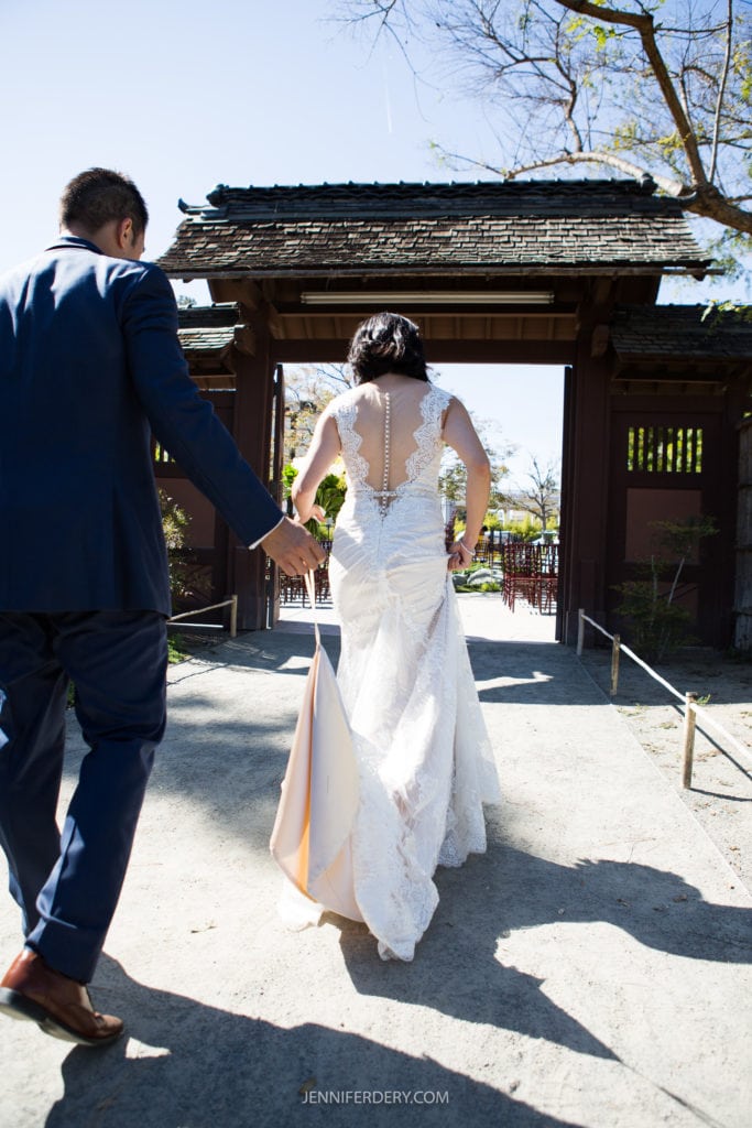 A bride and groom walk towards a traditional Japanese gate at their Japanese Friendship Garden wedding. The bride, in a white lace gown, is holding the groom's hand and lifting her dress slightly. The groom is wearing a navy blue suit. The background features trees and part of a garden.