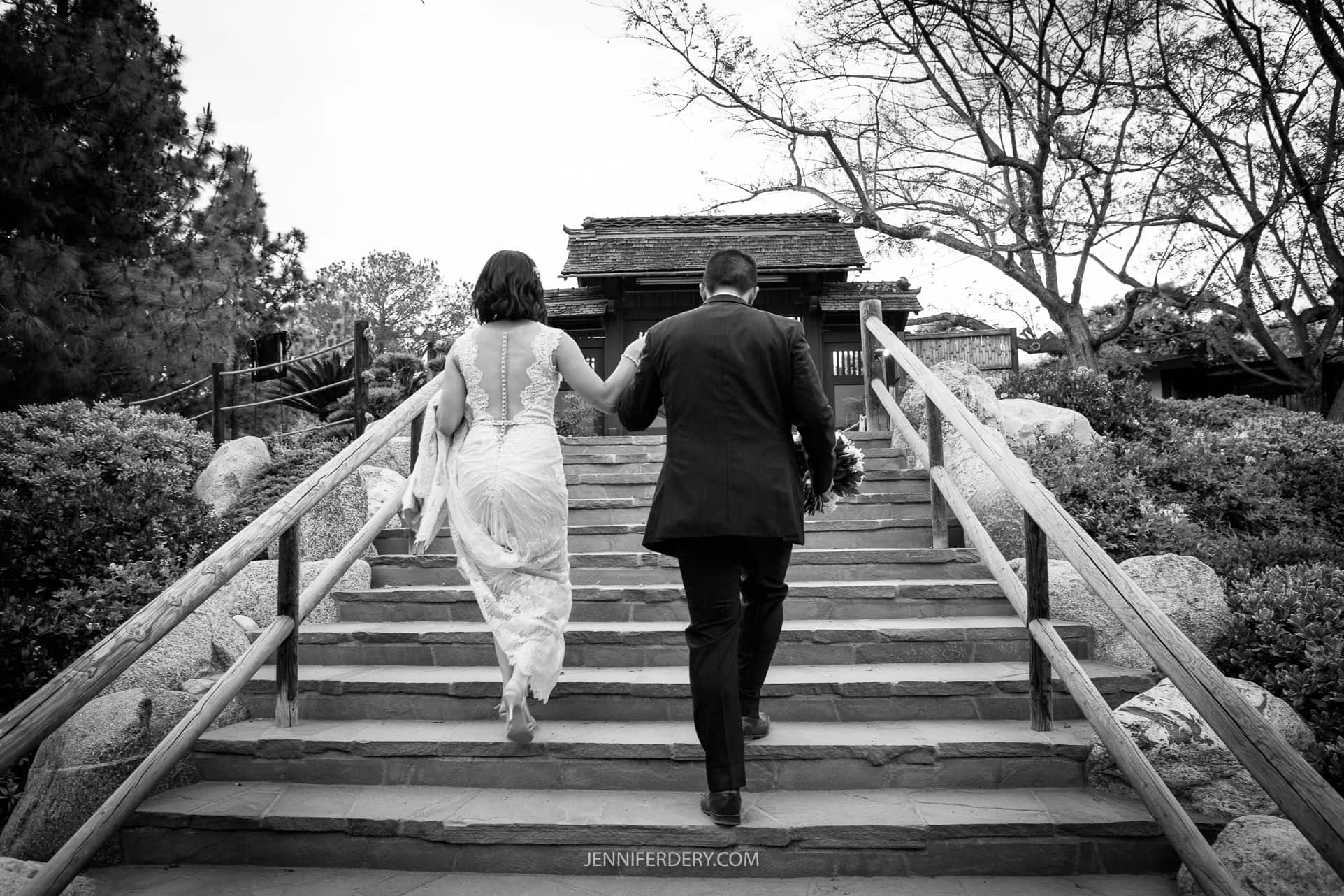 A black-and-white photo shows a couple walking up stone steps hand in hand. The woman on the left is in a lace wedding dress, and the man on the right is in a dark suit. They are ascending towards a building, surrounded by trees and shrubs, capturing a perfect Japanese Friendship Garden wedding moment.