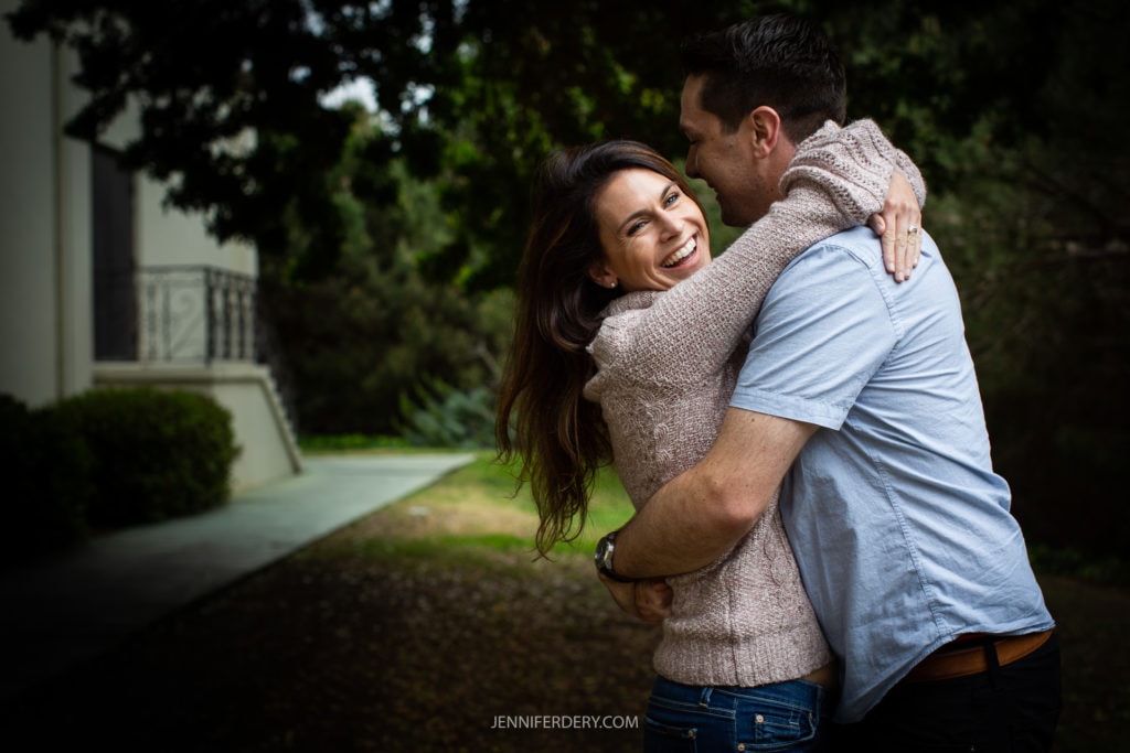 A couple is embracing and smiling outdoors on the campus of USD. The woman, wearing a pink sweater, is hugging the man, who is in a light blue shirt. They're standing on a lawn with trees and a building in the background.