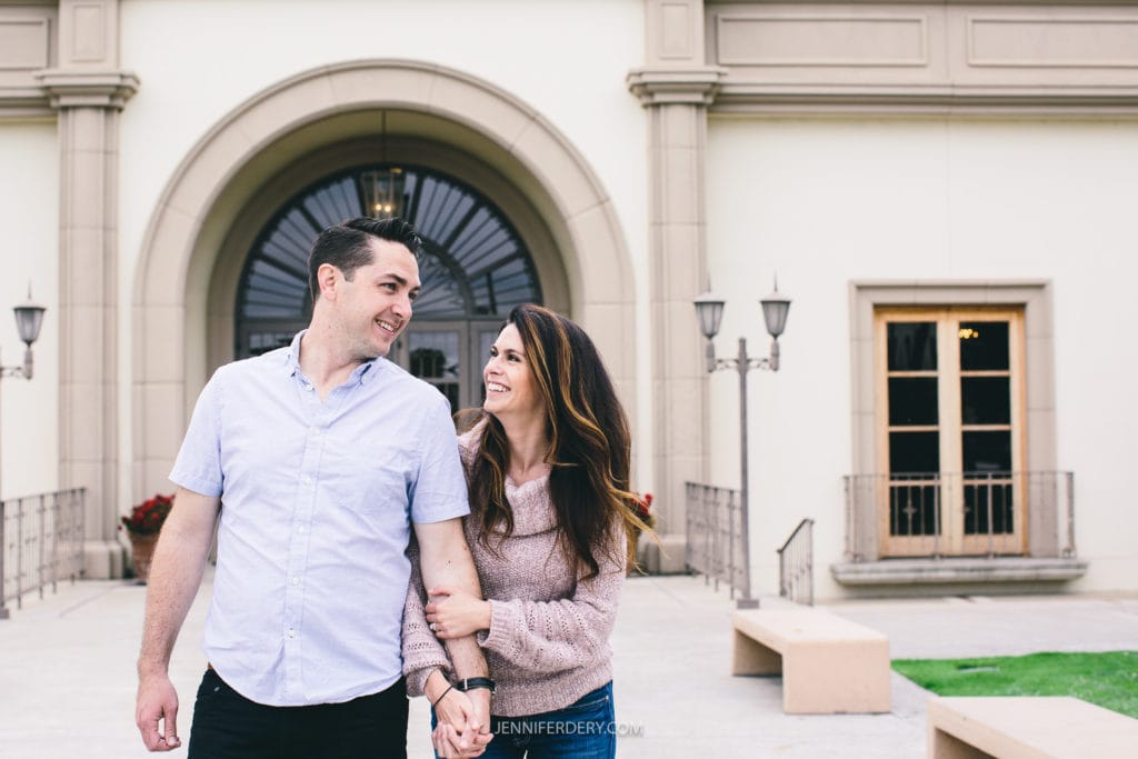 An engagement session at USD campus. A man and a woman are smiling at each other while walking outside a large building with arched windows and a grand entrance. The woman has long brown hair and is wearing a light pink sweater, and the man has short dark hair and is wearing a light blue shirt.
