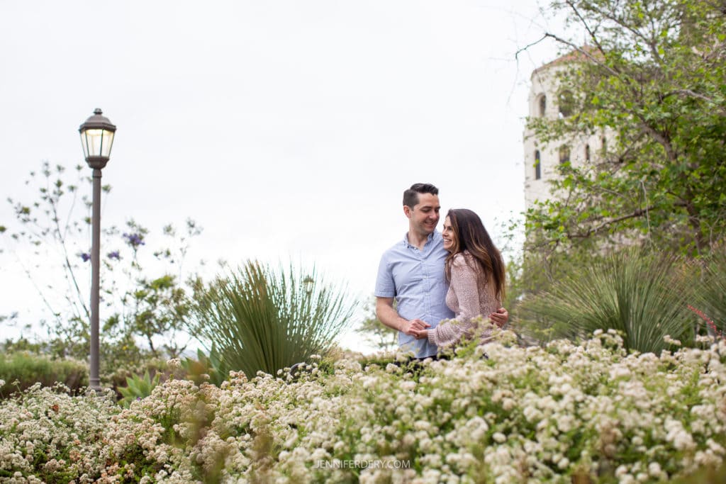 A couple stands closely, smiling at each other amidst lush greenery and white flowers at USD campus. They are surrounded by tall plants, with a vintage street lamp on the left and a rustic building partially visible on the right.